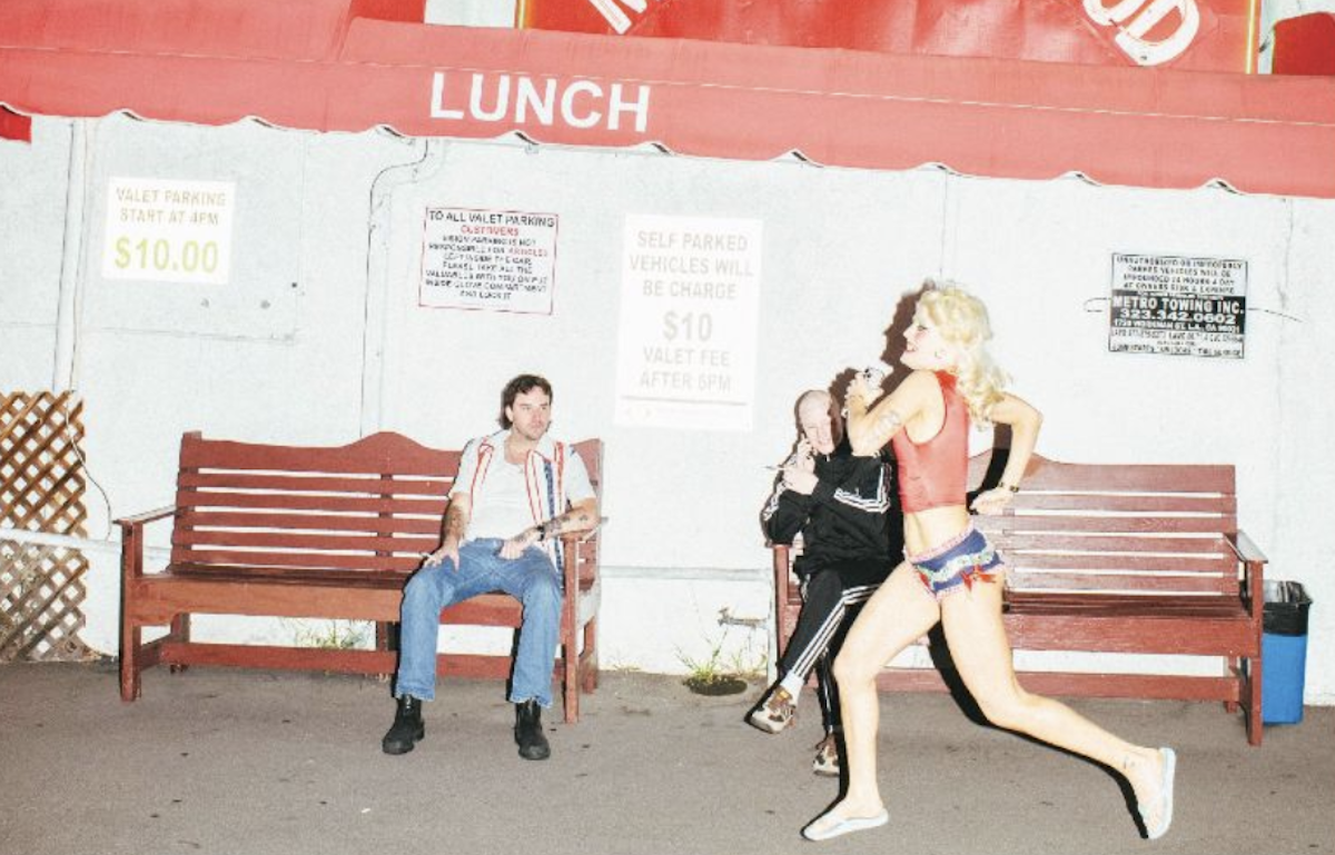 Punk band Amyl and the Sniffers outside a restaurant at night, featuring Amy Taylor running in a red crop top past bandmates sitting on wooden benches.